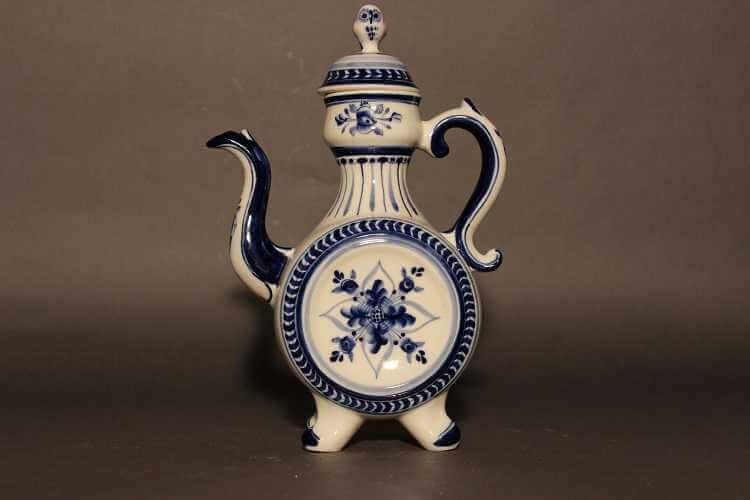 Tall blue and white coffeepot with curved spout, inverted S-scroll handle, and drum like body with blue floral motif on center of flat side and neck tapering inward and swelling slightly up to the lid with an owl finial.