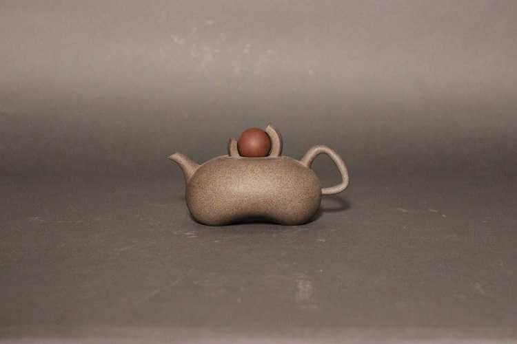 Speckled gray teapot with bean-shaped body, a very short spout and a circular inset lid with 2 curved pieces holding a loose red ball.