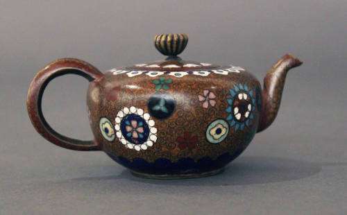 A dark brown colored teapot with a round handle, an outward curved spout, a cup-shaped body featuring circular floral designs, and a lid with ridged diamond-shaped finial.