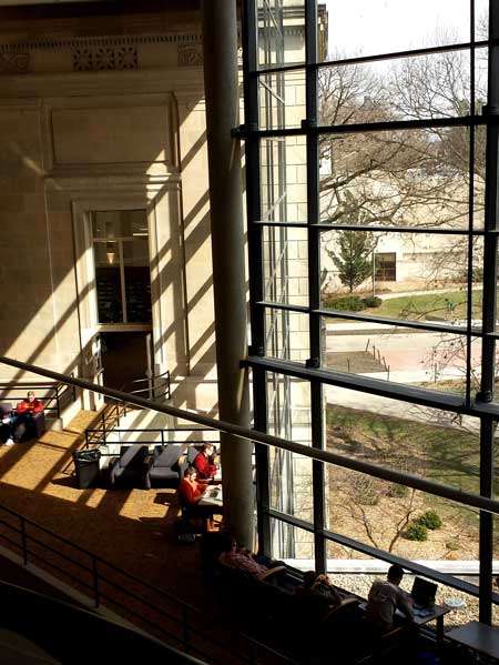 Photograph of a large windowed wall with sunlight peeking in and students seated by the wall taken from stairs in the library.