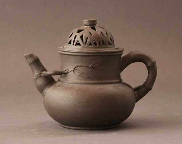 A blackish brown teapot with a flat bulbous bottom, a straight-sided neck, a bamboo-like handle and spout, and a lid pierced in a leaf pattern with a round finial.