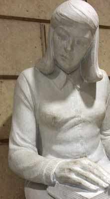 Sculpture of a seated girl reading a book on her lap looking slightly to her right.