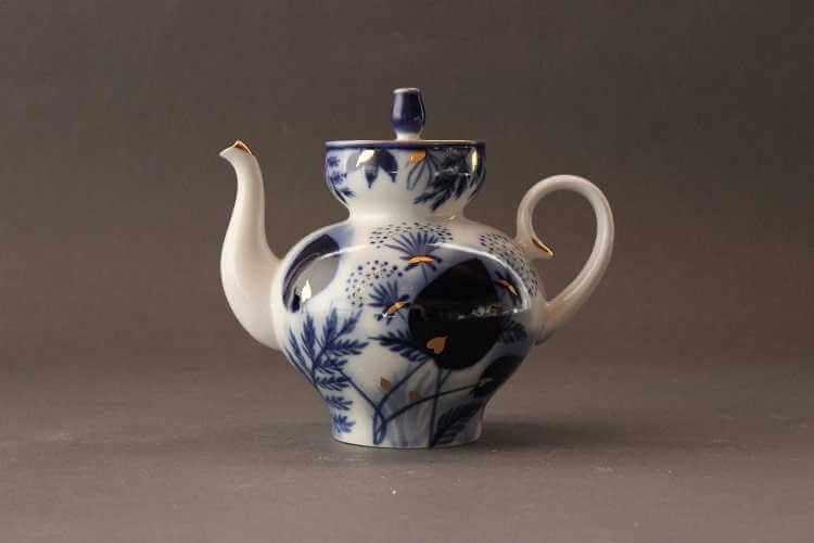 Blue and white teapot with narrow base and body swelling into a rounded shape featuring blue floral, leaf, and branch motif with extra shading and gilded accents. The neck is short with larger bowl-like area where the lid with tulip finial rests.