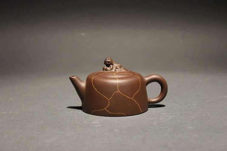 A brownish copper-colored teapot with a short straight spout and small, almost circular handle, a short squat-shaped body decorated with curving orangey brown lines to create a stone-like pattern, and a lid topped with a stylized dog of Fu finial.