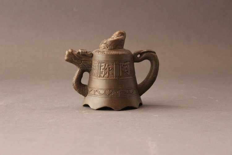 Teapot with a bell-shaped body covered in a thinner and thicker band of low relief work with Chinese characters and spout being the neck and head of a dragon and the handle being the tail. Lid finial is a laughing Buddha.