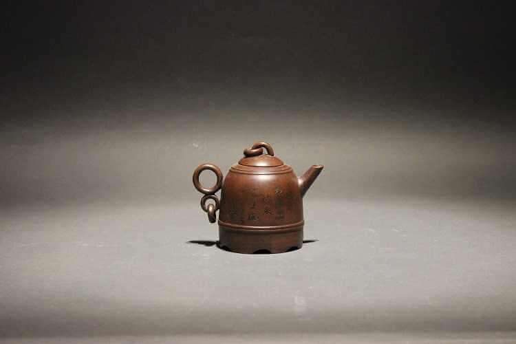 Dark reddish brown teapot with dome shaped body covered in incised Chinese characters and a handle that is a larger circle on top of a smaller circle with a loose ring hanging off from it.