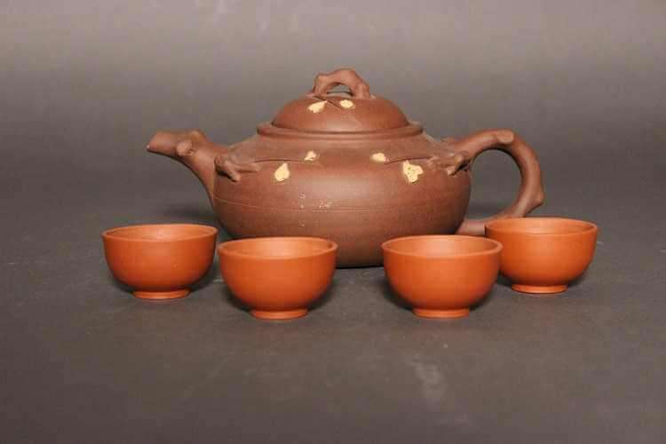 A brown teapot with a branch-like handle, spout, and finial, and a short and squat body with molded branch forms on the body along with yellow colored flowers. The teapot is surrounded by four small and plain redware cups without handles.