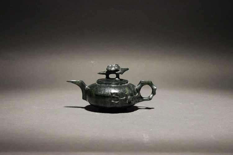 A dark green teapot with a squat circular body and a bamboo handle extending into a branch with a flower on the side of the teapot. There's a flower atop the triangular top of the handle on the lid.
