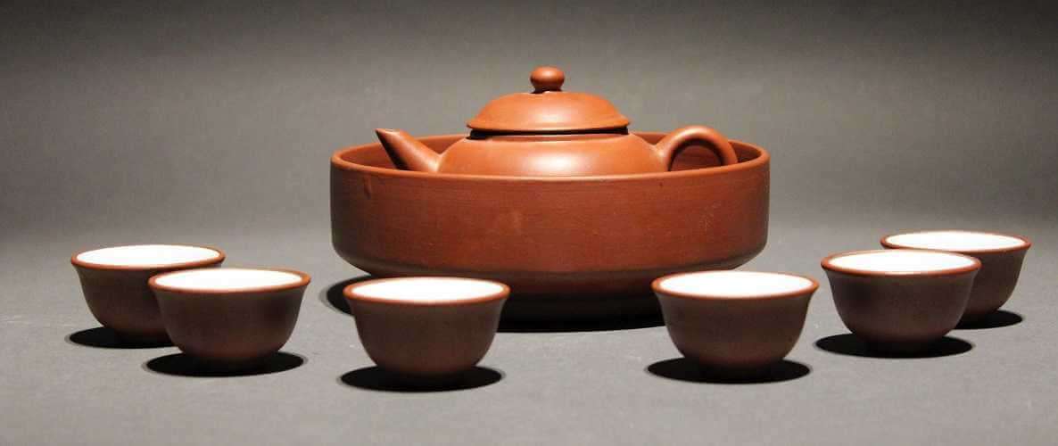 Brick red shallow bowl with straight sides holding a brick red teapot with a rounded body, straight spout and curved handle. Six cups that slightly flare at the lips surround the bowl.
