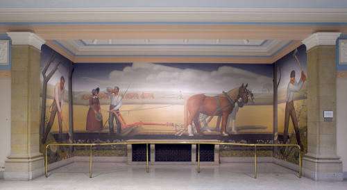 Wide Mural with a straight on view of a brown and a white horse with a red plow connected to them in a golden field under a blue sky with long fluffy clouds. A man in dark gray suspenders and a white t-shirt drinks water from a pitcher while standing next to the end of the plow. A woman in a blue bonnet and white-collared long-sleeve brown gown holds a hat and faces the man and an ox team breaks the prairie in the background. The mural on the left right angled wall shows a white man in long sleeve white t-s