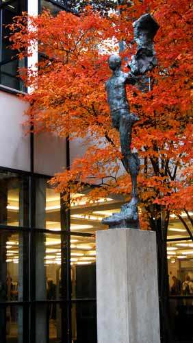 Sculpture of an angel with raised left wing but missing right limbs and wing, on a pedestal, taken from its left at an angle with a tree with orange colored leaves and windowed wall of the library in the background.
