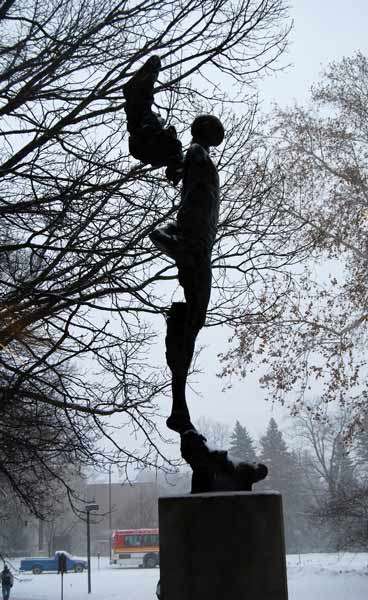 Sculpture of an angel with raised left wing but missing right limbs and wing, on a pedestal, taken from its right in black shadow with snowy ground and bare branches of trees in the background.