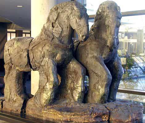 Sculpture of a pair of adjacent horses wearing harnesses. The horse on the left is ahead of the other and looks at the horse on the right.