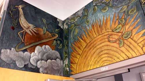 Murals above door near the corner of a wall. The mural on the left wall shows a crème bird sitting on a red planet with greenish red ring above the clouds against a deep blue backdrop. The mural on the right wall shows half a sun drawn with many rings and small spiky rays under leafy wines against a light centered blue backdrop.