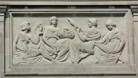 The bas-relief depicts four seated women. The first woman holds a paint palette in her right hand and a sculpture in her left hand while the second woman holds a fruit basket. The third woman holds a staff with yarn or thread wound around it and the fourth woman reads a scroll spread across her lap.