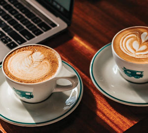 Open scholars graphic: Color photograph closeup of two cups of cappuccinos on saucers next to computer; all sitting on a wood surface.